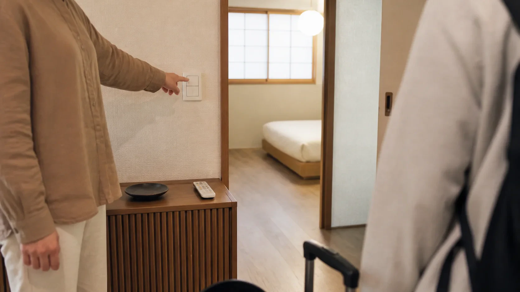 玄関から来客が入り、住人が客室の照明とリモコンの場所を示す場面